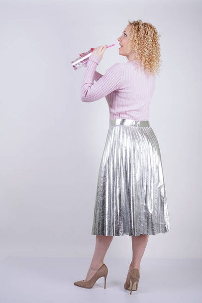 красивая счастливая белая женщина с вьющимися светлыми волосами стоит в тонком розовом трикотажном свитере и металлической серебряной складчатой длинной юбке с напитком в руках на белом фоне в Студии
 - Фото, изображение