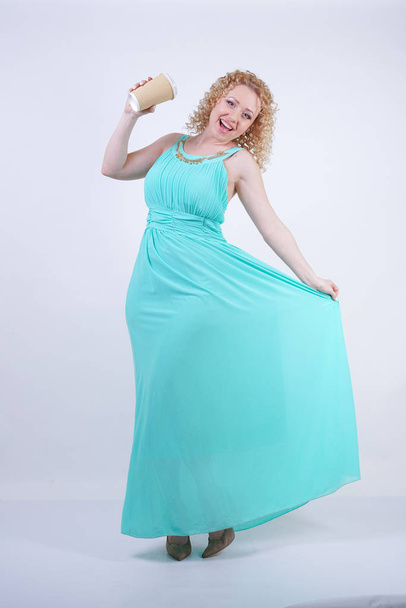 hübsche blonde kaukasische Frau trägt langes blaues Sommerkleid mit Pappbecher Kaffee und genießt das Leben auf weißem Studiohintergrund - Foto, Bild
