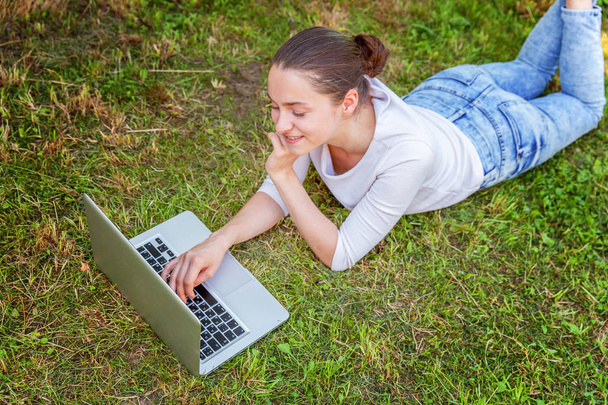 Young woman lying on green grass lawn in city park working on laptop pc computer. Freelance business concept - Photo, Image