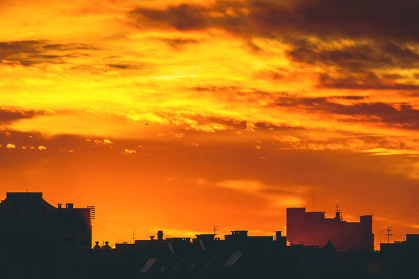 Cityscape с ярким огненным рассветом. Удивительное теплое драматическое облачное небо над темными силуэтами городских зданий. Оранжевый солнечный свет Атмосферный фон восхода солнца в пасмурную погоду. Копирование пространства
. - Фото, изображение