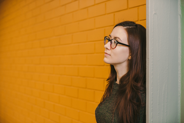 Beautiful cheerful pensive dreaming girl with long red hair dyed with henna, in green blouse and red glasses, on background of yellow brick wall, looks in sideways and away - Photo, Image