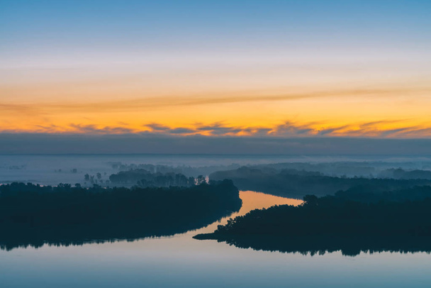 Early blue sky reflected in river water. Riverbank with forest under predawn sky. Yellow stripe in picturesque sky. Fog hid trees on island. Mystical morning atmospheric landscape of majestic nature. - Photo, Image