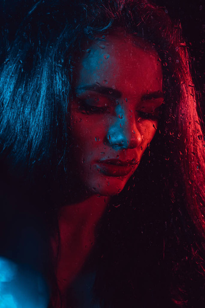 Retrato sensual de menina bonita atrás de vidro com gotas de chuva com iluminação azul vermelha
 - Foto, Imagem