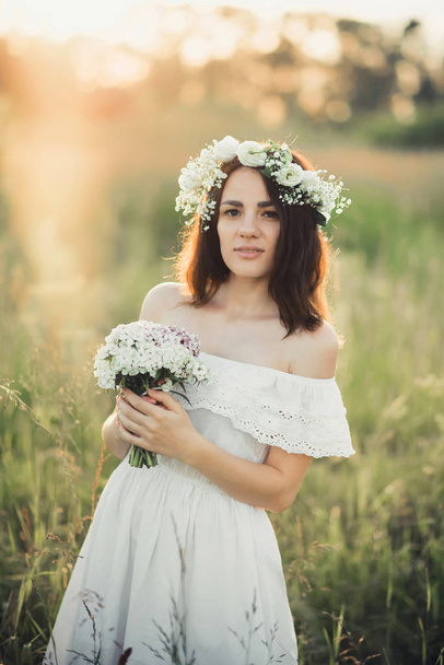 όμορφη νεαρή κοπέλα με ένα μπουκέτο από λουλούδια και ένα στεφάνι σε ένα λευκό φόρεμα - Φωτογραφία, εικόνα