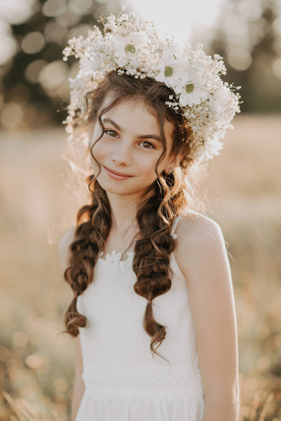 πορτρέτο ενός χαμογελαστή νεαρό κορίτσι με ένα λευκό φόρεμα και ένα λουλουδάτο στεφάνι στα μαλλιά της με πλεξούδες στο γήπεδο του καλοκαιριού - Φωτογραφία, εικόνα