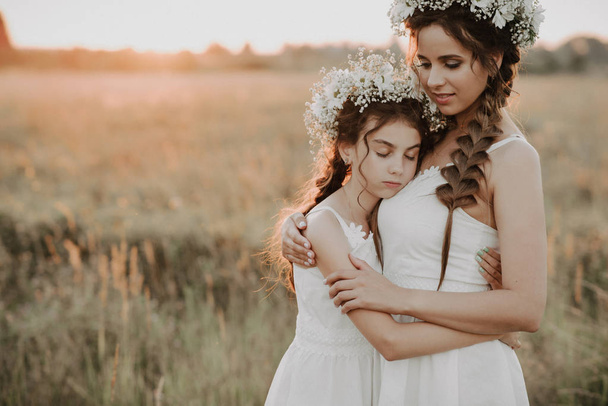 mother and daughter together in white dresses with braids and floral wreaths in boho style in the summer field at sunset - Foto, imagen