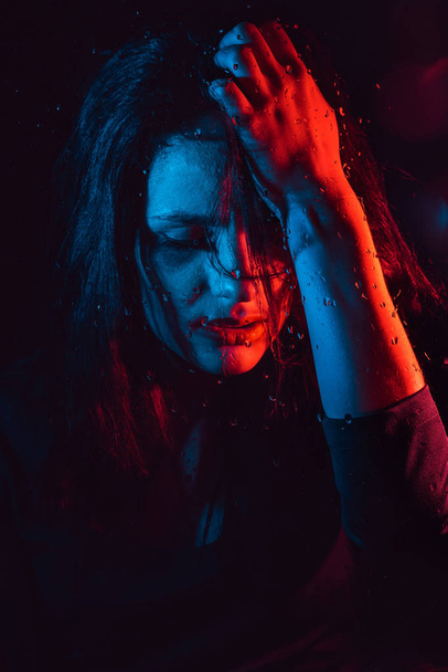 Sensual portrait of beautiful girl behind glass with raindrops with red blue lighting - Foto, Imagen