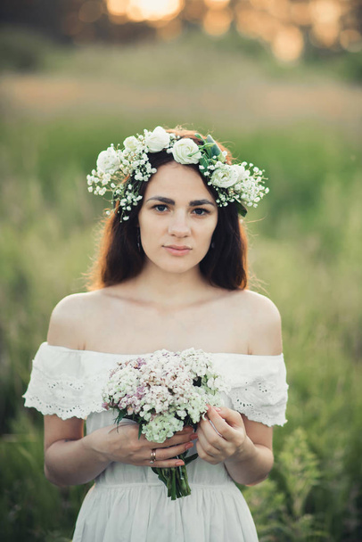 portrait of an attractive girl in white dress with a bouquet of flowers and a wreath in summer - Foto, Imagen