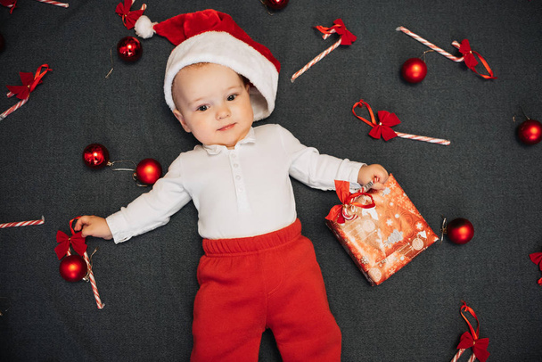 Malý chlapeček v Santa Klause leží mezi vánočními bonbony, míčky a dárkovým políčkem - Fotografie, Obrázek