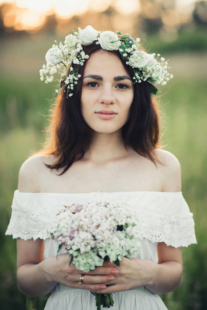 portrait of an attractive girl in white dress with a bouquet of flowers and a wreath in summer - Фото, изображение