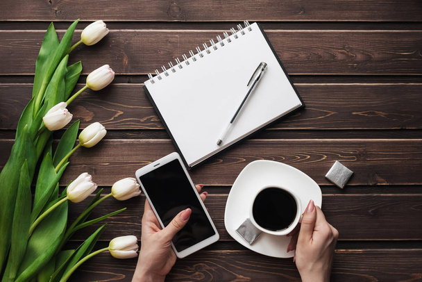 White tulips on a wooden table with an empty notebook, smartphone and a Cup of coffee in women's hands - Photo, Image