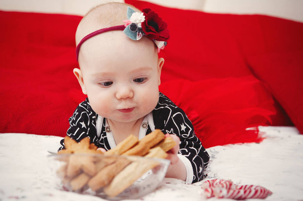 Красива п'ятимісячна дівчинка-малюк з квіткою на голові в блакитній сукні з різдвяними пряниками на червоному тлі. Догляд за мамою. Догляд за дітьми. Крупним планом
. - Фото, зображення