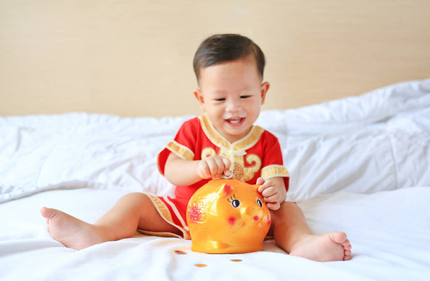 Smiling little Asian baby boy in traditional Chinese dress putting some coins into a piggy bank sitting on bed at home. Kid saving money concept. Focus at piggybank. - Фото, зображення