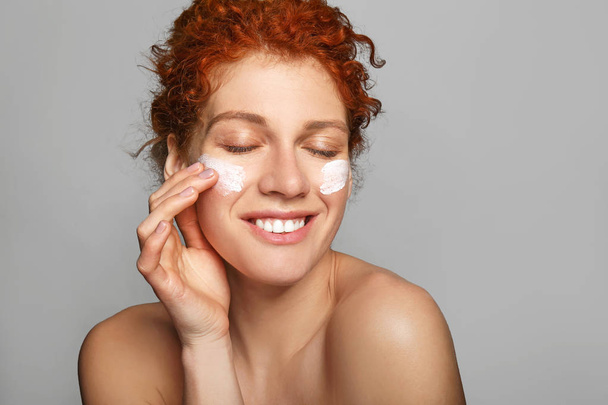 Beautiful redhead woman applying facial cream against grey background - 写真・画像