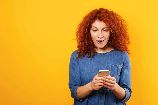 Surprised redhead woman with mobile phone on color background - Фото, изображение