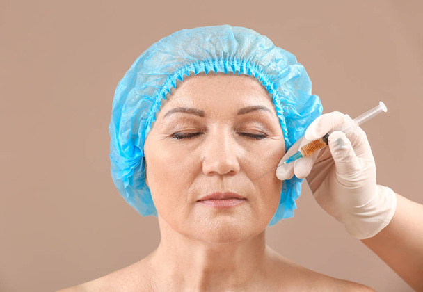 Mature woman and hand of plastic surgeon with syringe on color background - Photo, image