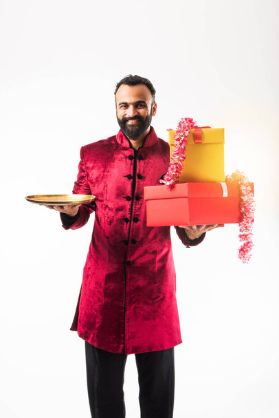 Ινδικό άνθρωπος κρατώντας στεφάνι λουλουδιών, κουτιά δώρων και άδεια πλάκα στο Σάλι/γάμο ή φεστιβάλ, φορώντας παραδοσιακά υφάσματα/Σεράνι, στέκεται απομονωμένη σε λευκό φόντο - Φωτογραφία, εικόνα