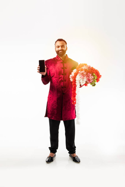Индийский мужчина, держащий гирлянду / серию цветов для украшения на дивали / свадьба или фестивали во время ношения традиционных тканей / шервани, стоя изолированы на белом фоне
 - Фото, изображение