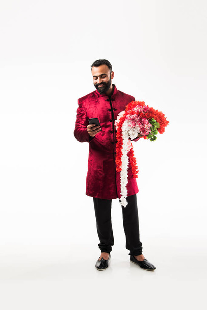 Ινδικό άνθρωπος κρατώντας λουλούδι στεφάνι/σειρά για τη διακόσμηση στο παάλι/γάμο ή φεστιβάλ, φορώντας παραδοσιακά υφάσματα/Σεράνι, στέκεται απομονωμένη σε λευκό φόντο - Φωτογραφία, εικόνα