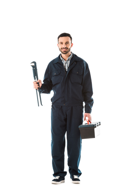 happy plumber in overalls holding toolbox and adjustable wrench while smiling at camera isolated on white - Foto, Bild