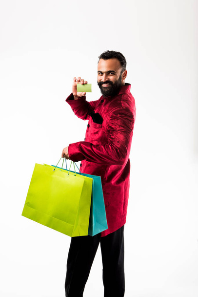 Indian man showing Card while shopping in red traditional wear, standing isolated with colourful paper bags over white background - Zdjęcie, obraz