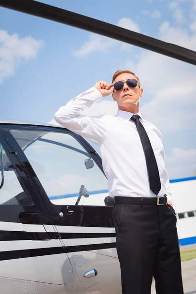 Pilot in formal wear adjusting sunglasses near helicopter - Photo, Image