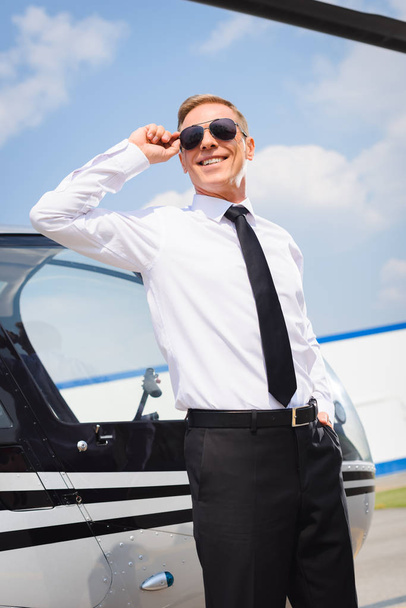 pilota sorridente in abbigliamento formale regolazione occhiali da sole vicino elicottero
 - Foto, immagini