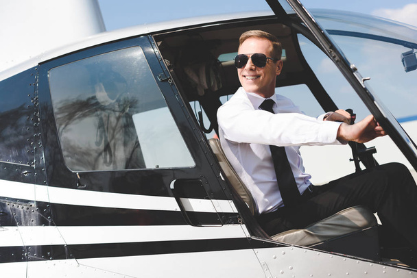Piloto guapo en ropa formal y gafas de sol sentado en cabina de helicóptero
 - Foto, Imagen