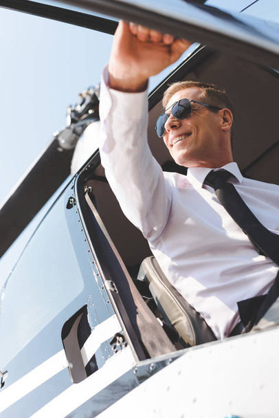 smiling Pilot in sunglasses and formal wear sitting in helicopter cabin and opening door - Valokuva, kuva