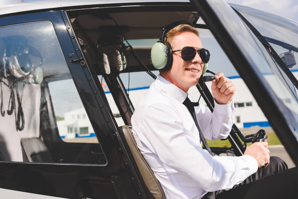 mosolyogva pilóta napszemüveg és headset ül helikopter kabin - Fotó, kép