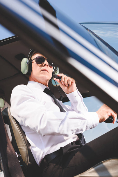 vista de bajo ángulo del piloto en gafas de sol y auriculares sentados en cabina de helicóptero
 - Foto, Imagen