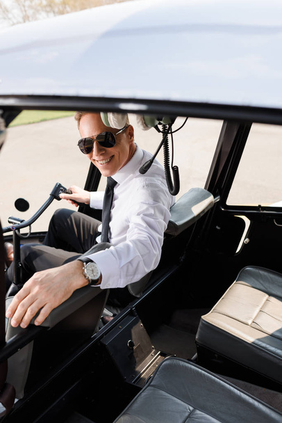 usměvavý pilot ve formálním opotřebení a slunečních brýlích sedícího v kabině vrtulníků - Fotografie, Obrázek