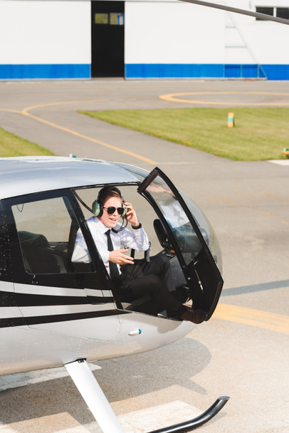Piloto maduro en ropa formal y gafas de sol sentado en cabina de helicóptero con puerta abierta
 - Foto, Imagen