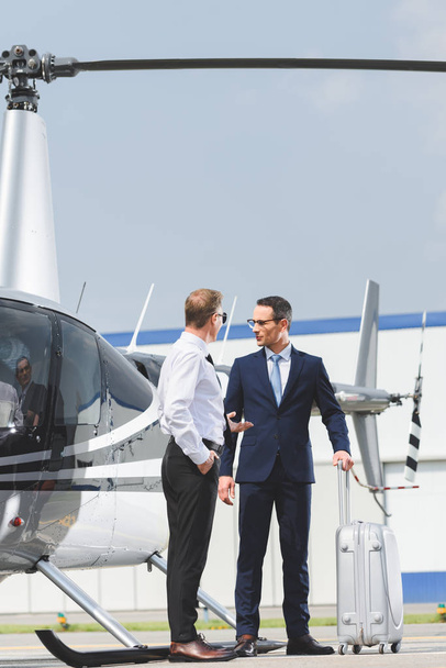 Piloto en ropa formal y hombre de negocios con maleta cerca de helicóptero
 - Foto, imagen