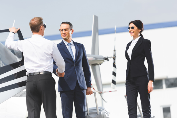 businessman with luggage shaking hands with pilot near helicopter and businesswoman - Foto, Imagen
