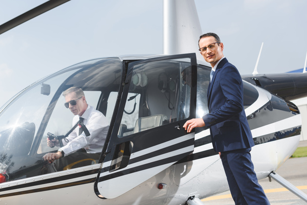 Piloto en ropa formal sentado en helicóptero mientras hombre de negocios mirando a la cámara
 - Foto, imagen