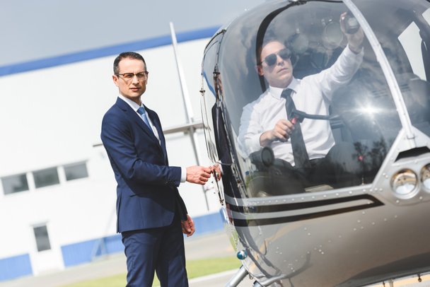 Пилот в формальной одежде сидит в вертолете, пока бизнесмен смотрит в камеру
 - Фото, изображение