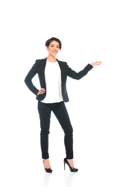 smiling mixed race businesswoman holding hand on hip and gesturing while looking at camera isolated on white - Foto, Bild