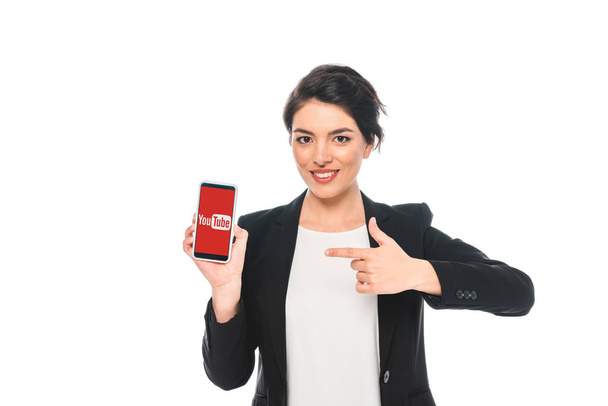 KYIV, UKRAINE - APRIL 24, 2019: Attractive mixed race businesswoman pointing with finger at smartphone with Youtube app on screen isolated on white. - Fotografie, Obrázek