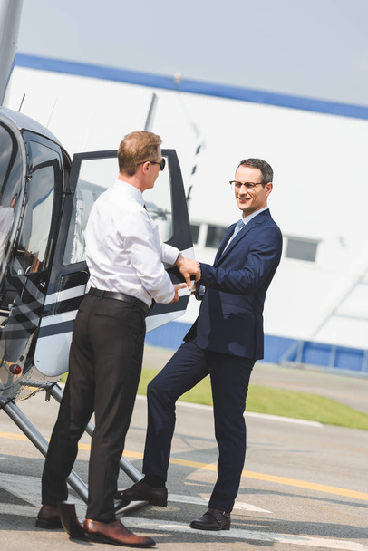 Piloto en ropa formal cerca de hombre de negocios abriendo la puerta del helicóptero
 - Foto, Imagen