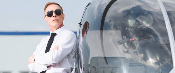 tiro panorâmico de piloto em óculos de sol e desgaste formal com braços cruzados perto de helicóptero
 - Foto, Imagem