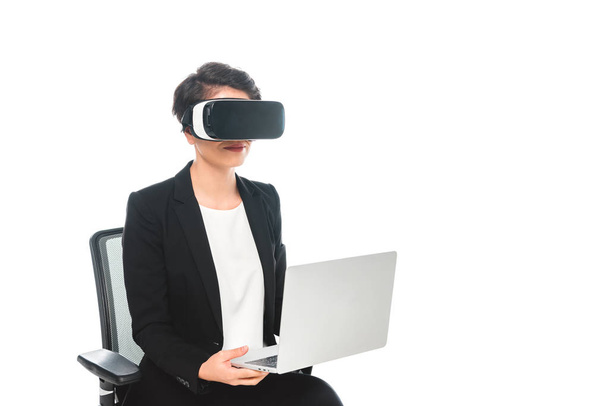 junge gemischte Rasse Geschäftsfrau in Virtual-Reality-Headset sitzt im Bürostuhl und mit Laptop isoliert auf weiß - Foto, Bild