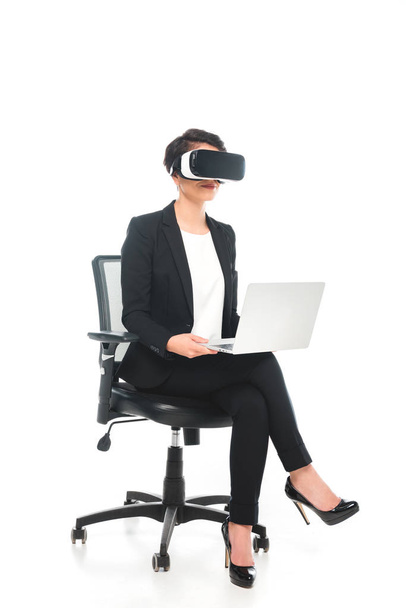 mladá smíšená závod podnikatelka ve virtuální realitě headset pomocí notebooku, zatímco sedí v kancelářské židli na bílém pozadí - Fotografie, Obrázek