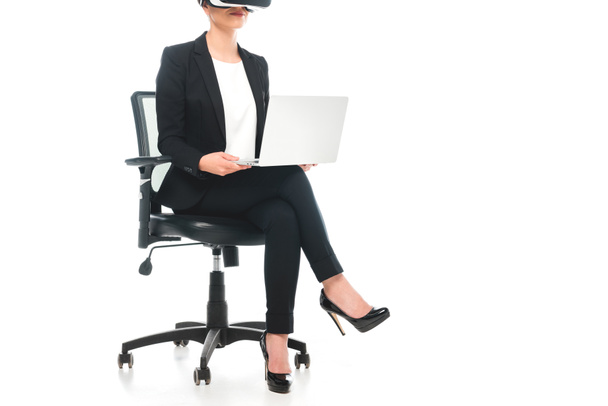 Teilansicht einer gemischten Geschäftsfrau mit Virtual-Reality-Headset, während sie im Bürostuhl sitzt und Laptop auf weißem Hintergrund benutzt - Foto, Bild