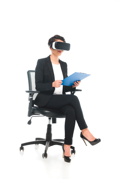 mujer de negocios de raza mixta usando auriculares de realidad virtual mientras está sentado en la silla de oficina y sujetando el portapapeles sobre fondo blanco
 - Foto, imagen