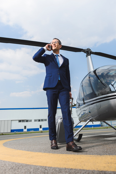businessman in formal wear with suitcase talking on smartphone near helicopter - 写真・画像