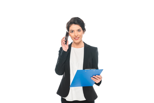 atractiva mujer de negocios de raza mixta hablando en el teléfono inteligente mientras sostiene el portapapeles y mirando a la cámara aislada en blanco
 - Foto, imagen