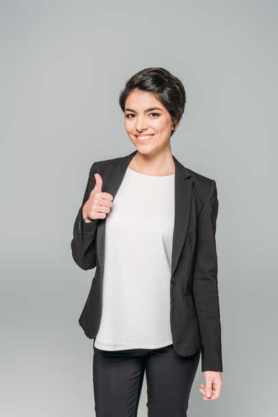 gelukkige gemengde race zakenvrouw die duim omhoog en glimlachend op de camera geïsoleerd op grijs - Foto, afbeelding