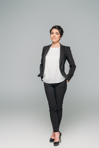 smiling mixed race businesswoman holding hands in pockets and looking at camera on grey background - Photo, Image
