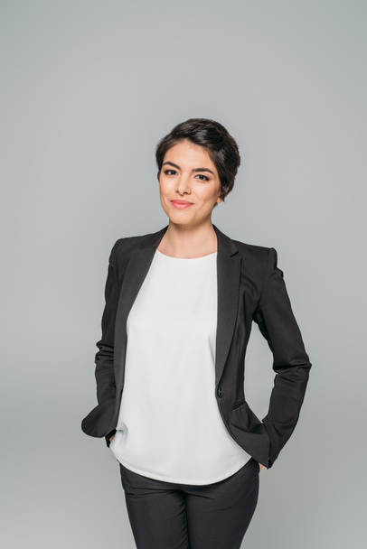 pretty mixed race businesswoman standing with hands in pocket and smiling at camera isolated on grey - Zdjęcie, obraz